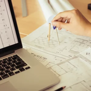 Architect working on blueprint in workplace with laptop and drawing compass.